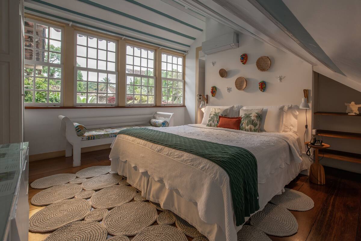 A bedroom with a large bed, a bench, and a view of greenery through the windows.