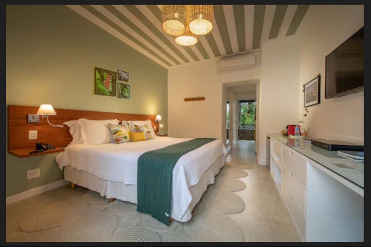A hotel room with a bed, bedside table, lamp, artwork on the wall, and a kitchenette area.