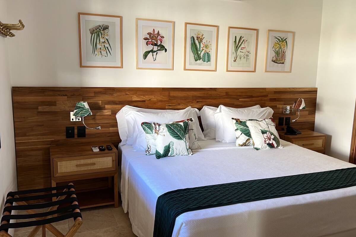 A hotel room with a wooden headboard, a bed with white bedding and tropical-themed pillows, a bedside table with a lamp, and framed botanical prints on the wall.