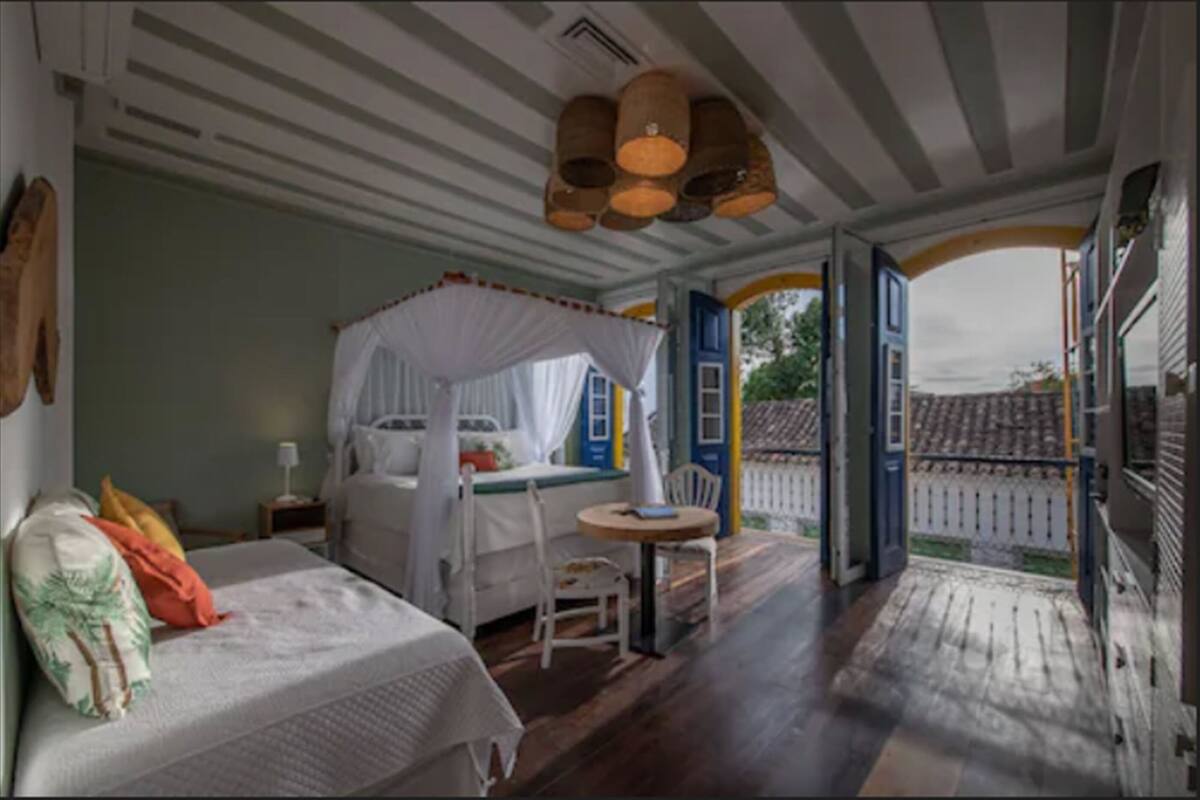 A bedroom with a four-poster bed, a small round table, and a chair. There is a window with a view of the outside.