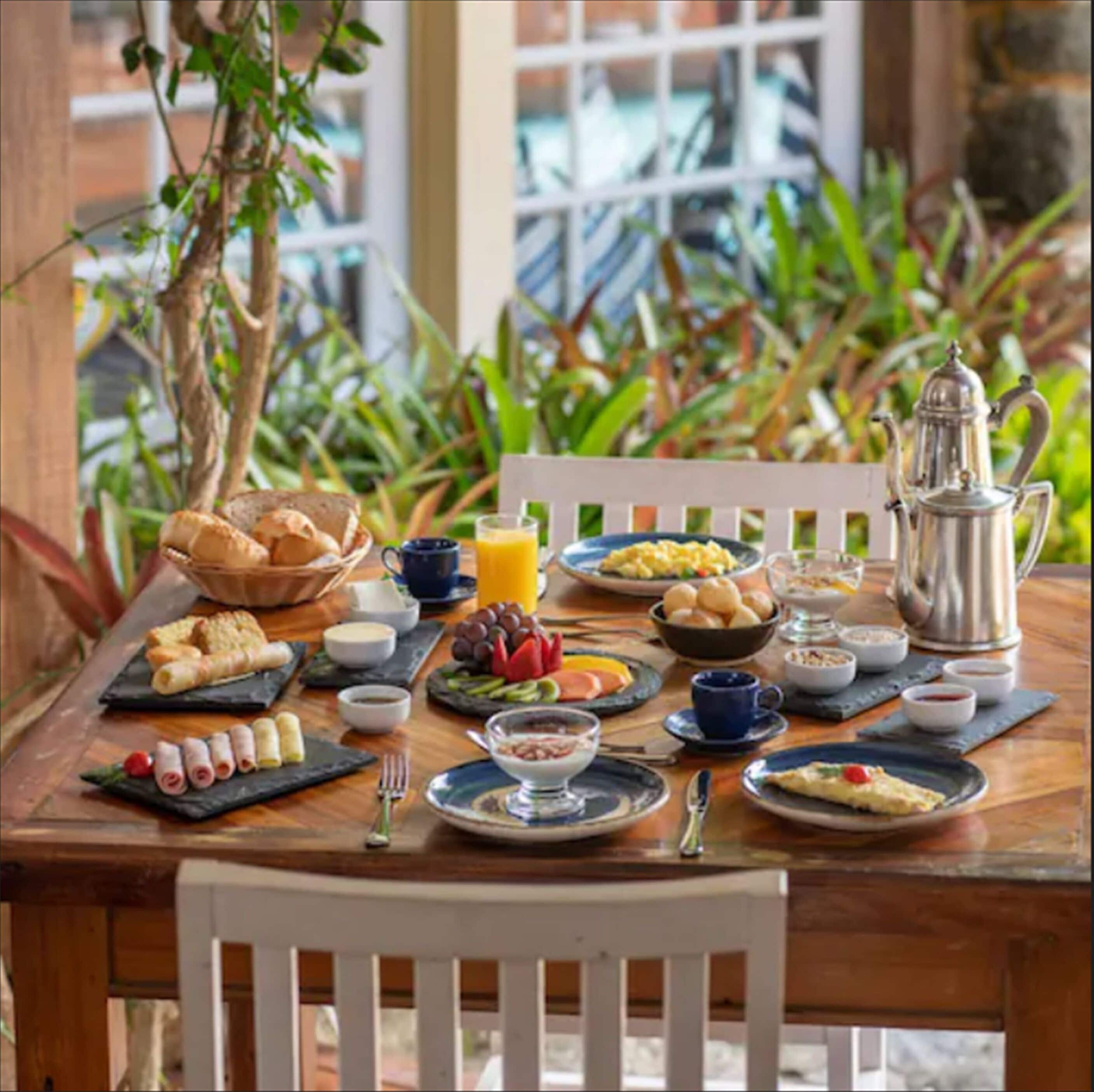 A breakfast table set with various dishes, including eggs, toast, and fruit, with a coffee pot and a view of greenery outside.