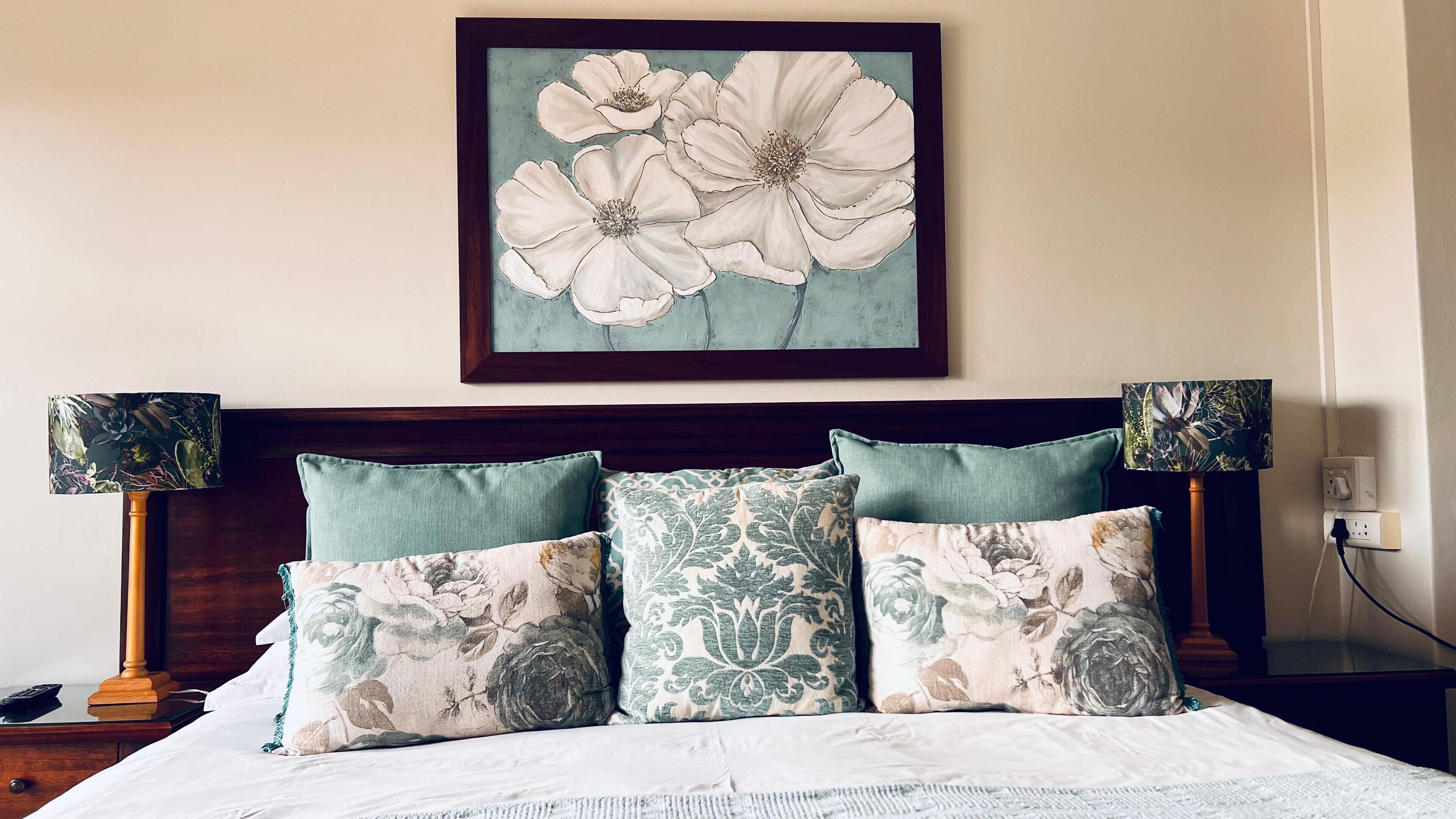 Een net opgemaakt bed met bloemenkussens en een bloemig kunstwerk erboven.