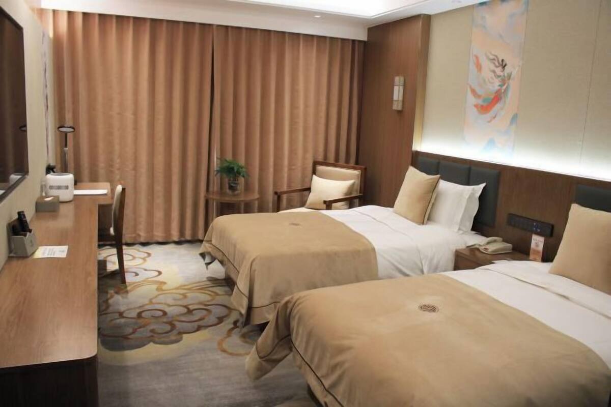 A hotel room with two beds, a desk, a chair, a small table with a plant, and a painting on the wall.