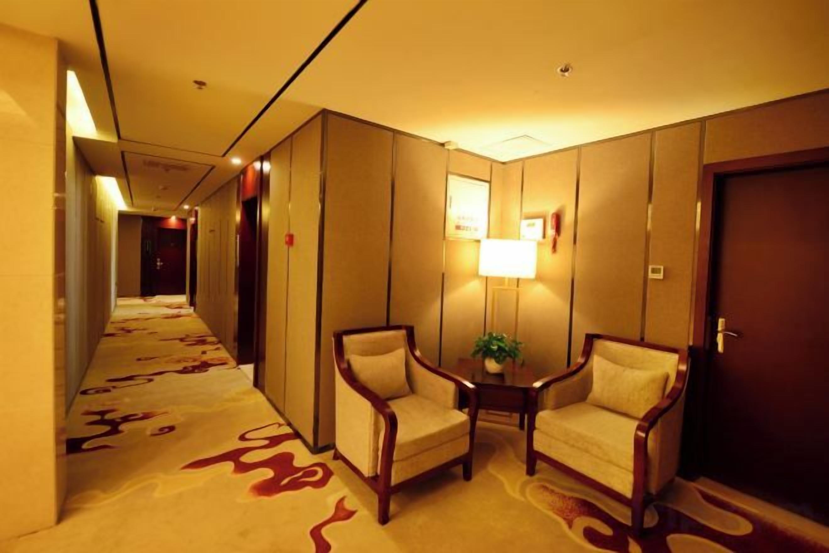A hotel hallway with carpeted floors, a reception desk, two armchairs, a small table with a plant, and a door on the right.