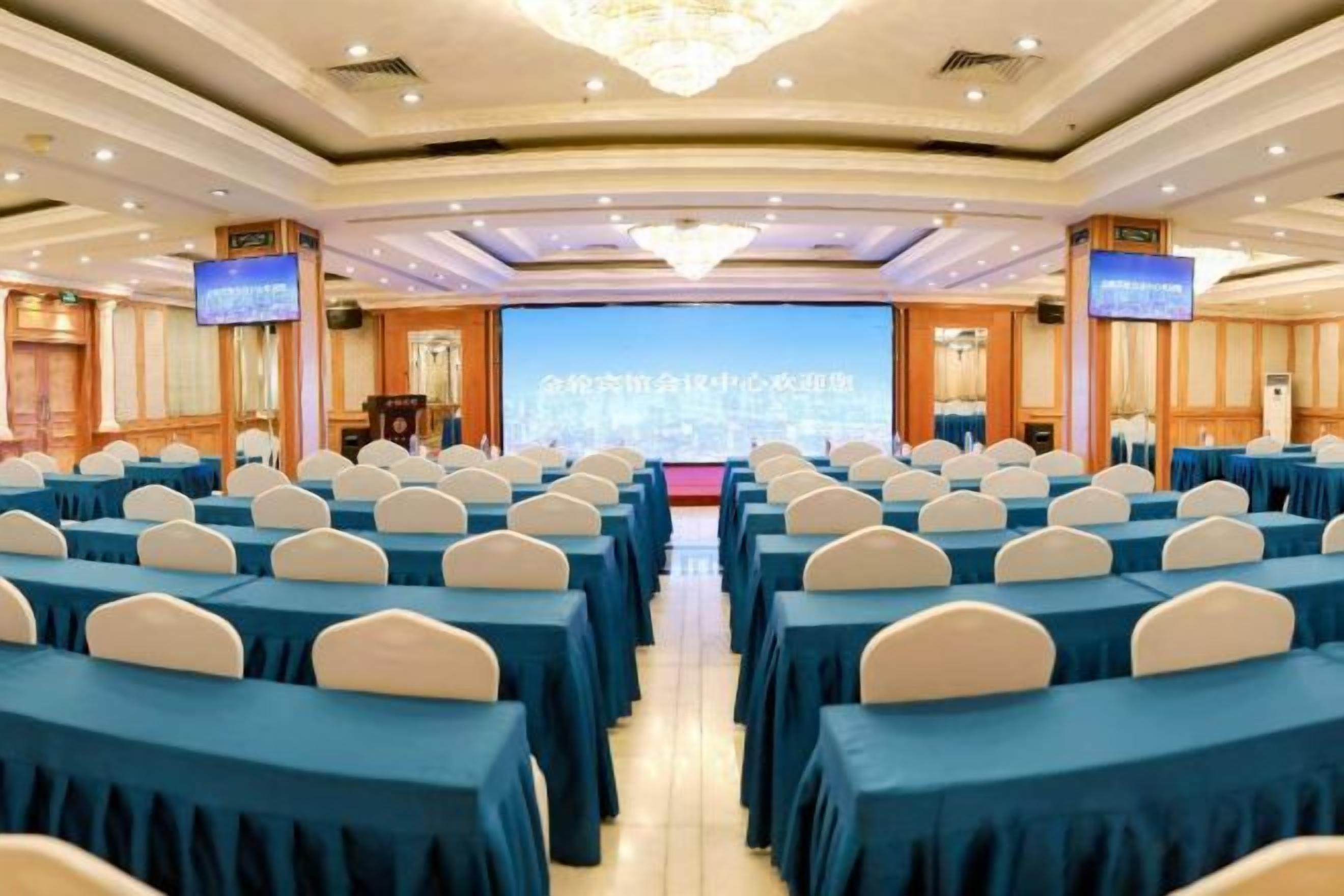 Une salle de conférence avec des rangées de tables et de chaises, un grand écran à l’avant et un lustre.
