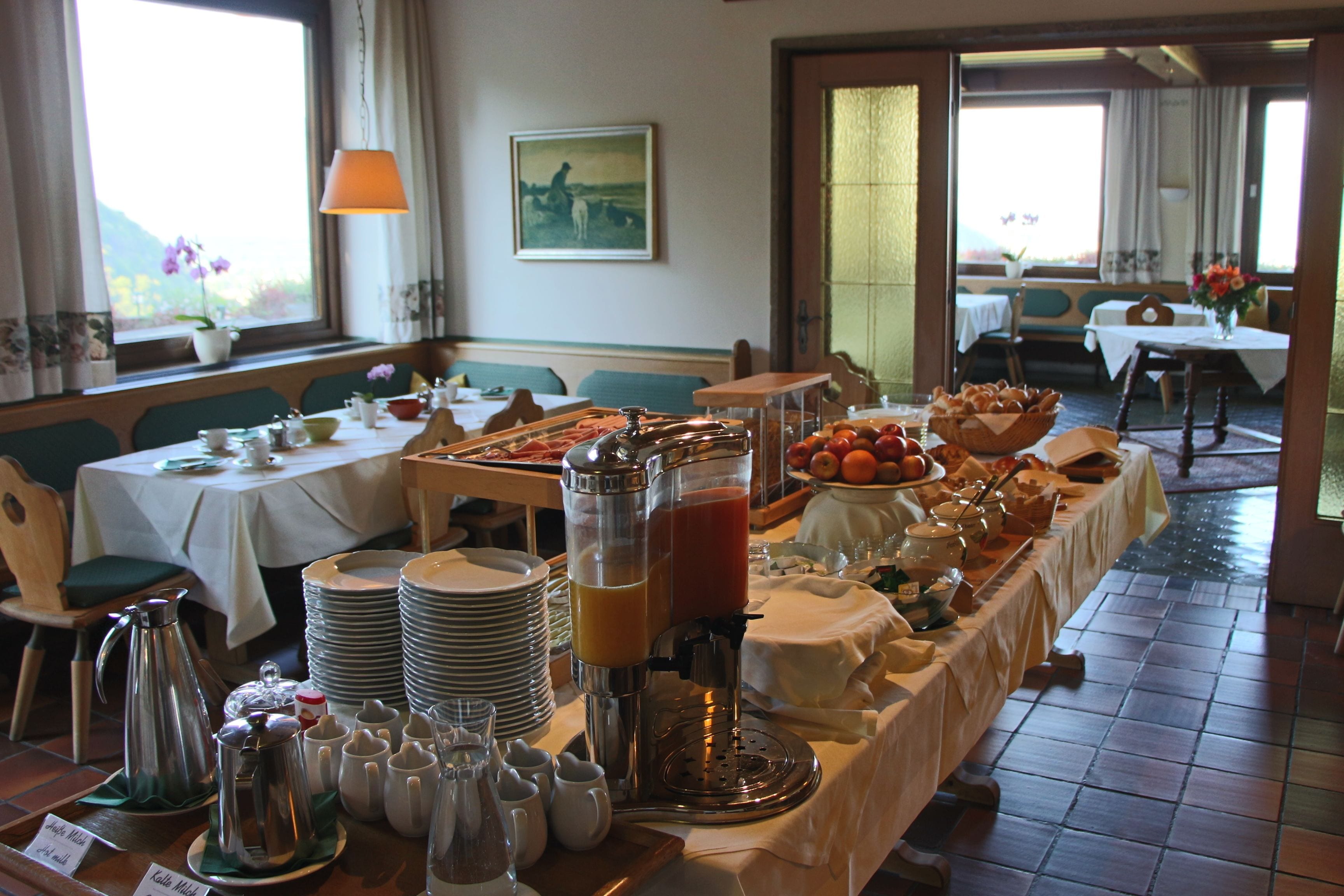 A breakfast buffet with various food items and beverages, tables with chairs, and a view of the outdoors through large windows.