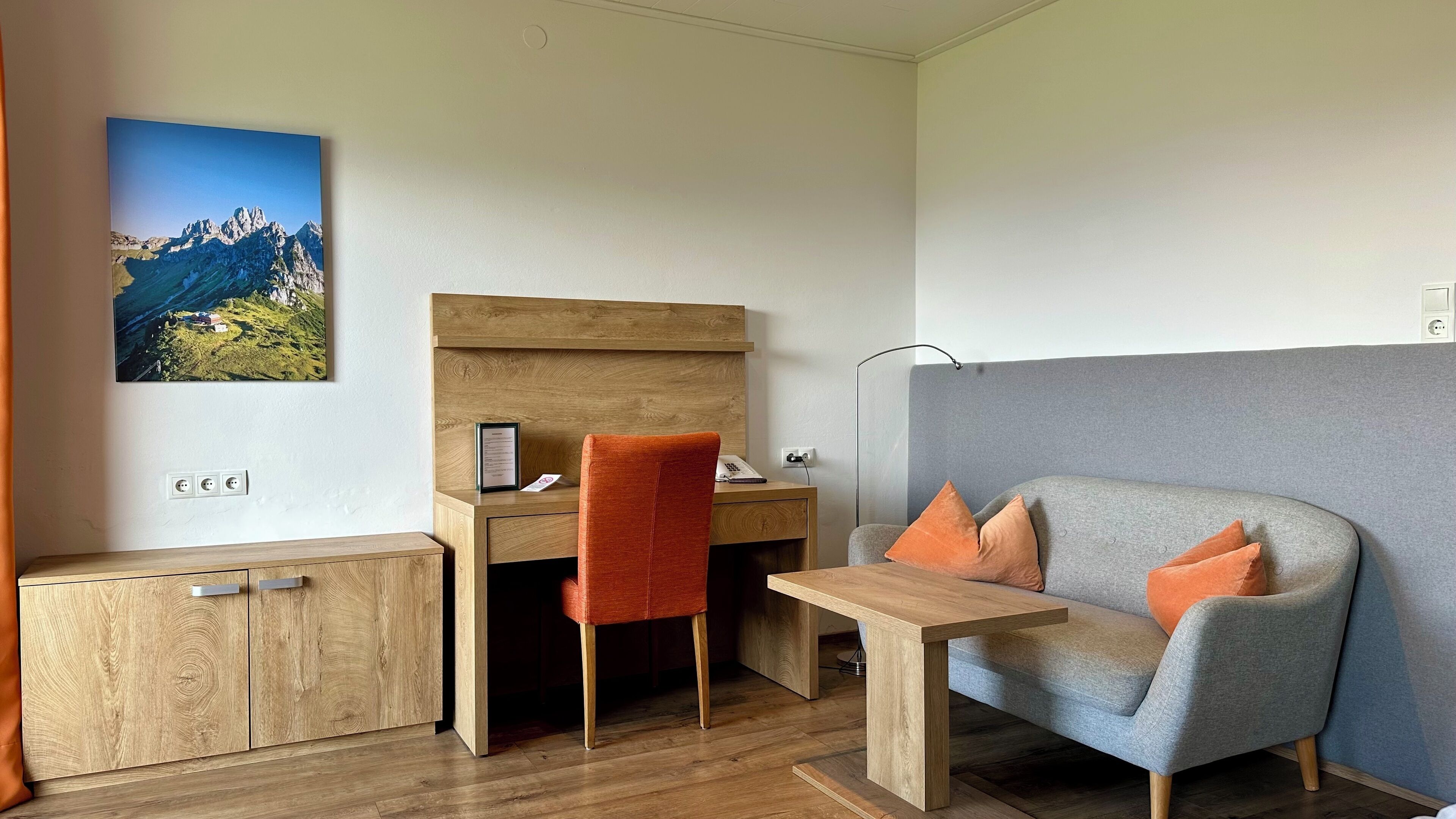Habitación moderna con un escritorio de madera, un sofá gris con cojines naranjas, una silla roja y una pintura con vistas a la montaña.