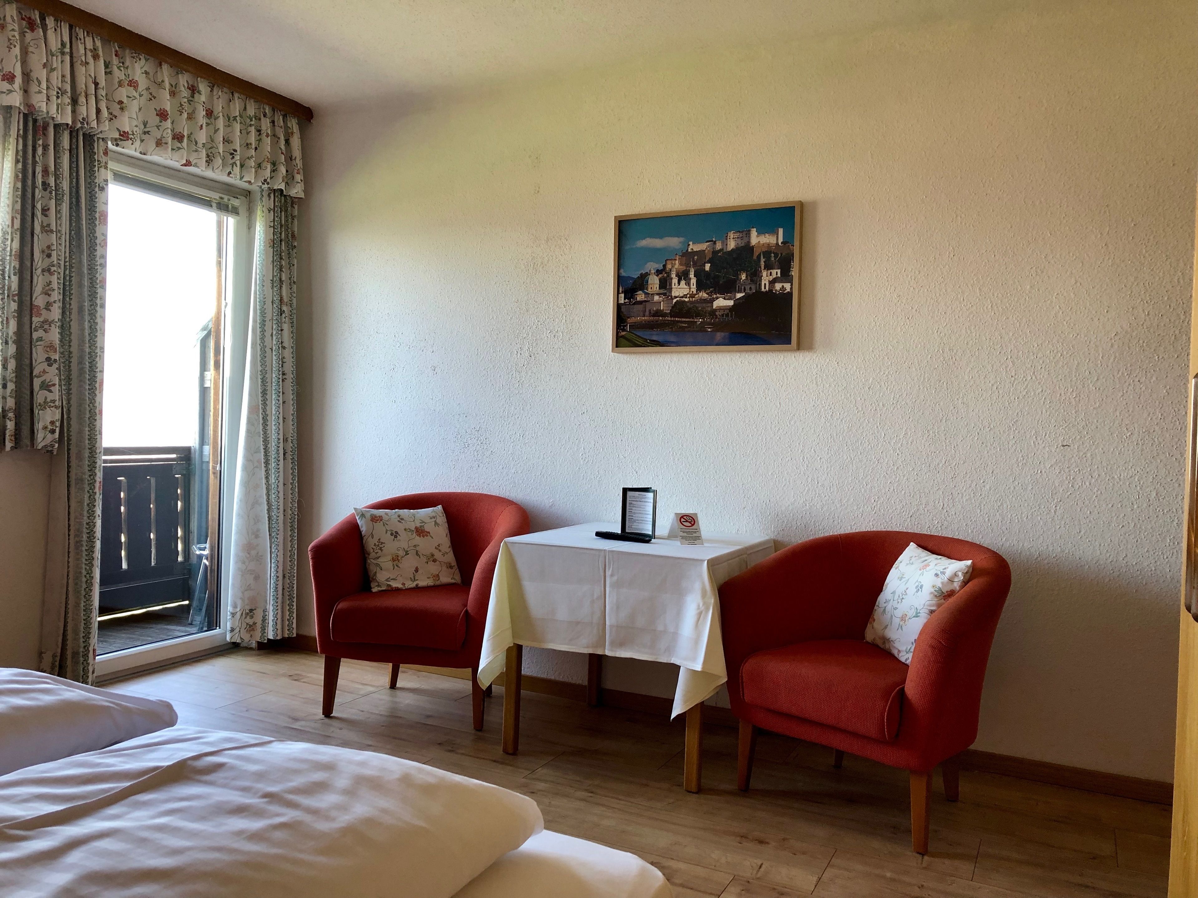 A hotel room with a bed, two red armchairs, a small table with a menu, and a cityscape painting on the wall.
