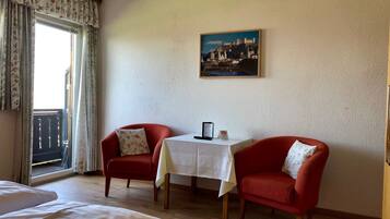 A hotel room with a bed, two red armchairs, a small table with a menu, and a cityscape painting on the wall.