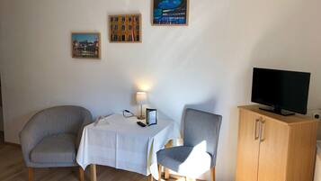 Ett litet rum med två randiga fåtöljer, ett bord med ett vitt bordsduksöverdrag, en lampa, en tv på ett träskåp och tre inramade tavlor på väggen.