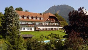 A large building with multiple windows and a sloped roof, surrounded by greenery and a mountain in the background.