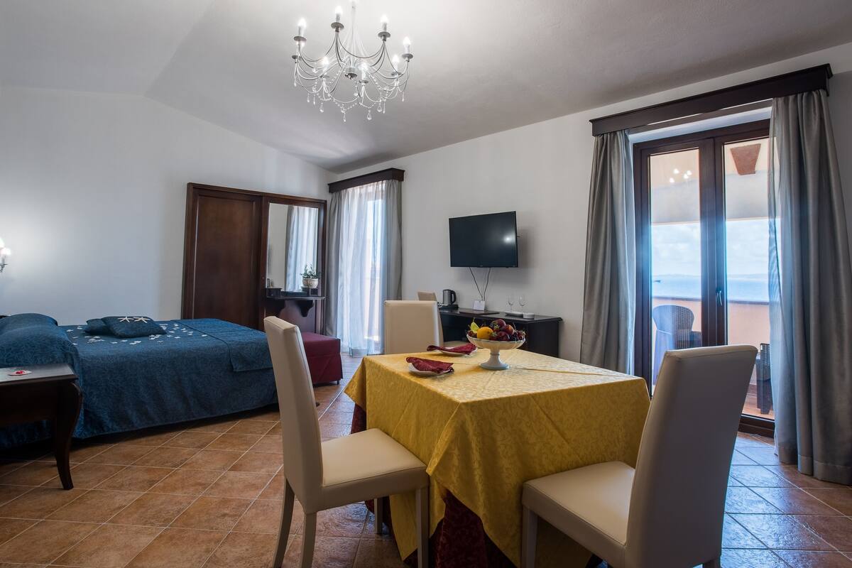 Ein Hotelzimmer mit einem Bett, einem Esstisch mit Stühlen, einem Fernseher und einem Balkon mit Vorhängen.