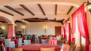 Un comedor con mesas redondas cubiertas con manteles rojos, sillas tapizadas en azul y vigas de madera en el techo.