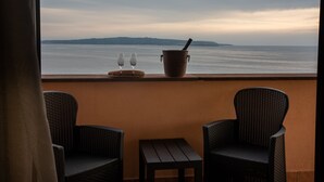 Un balcón con dos sillas de mimbre, una mesita y dos vasos sobre una bandeja, con vistas a un mar tranquilo y colinas lejanas.