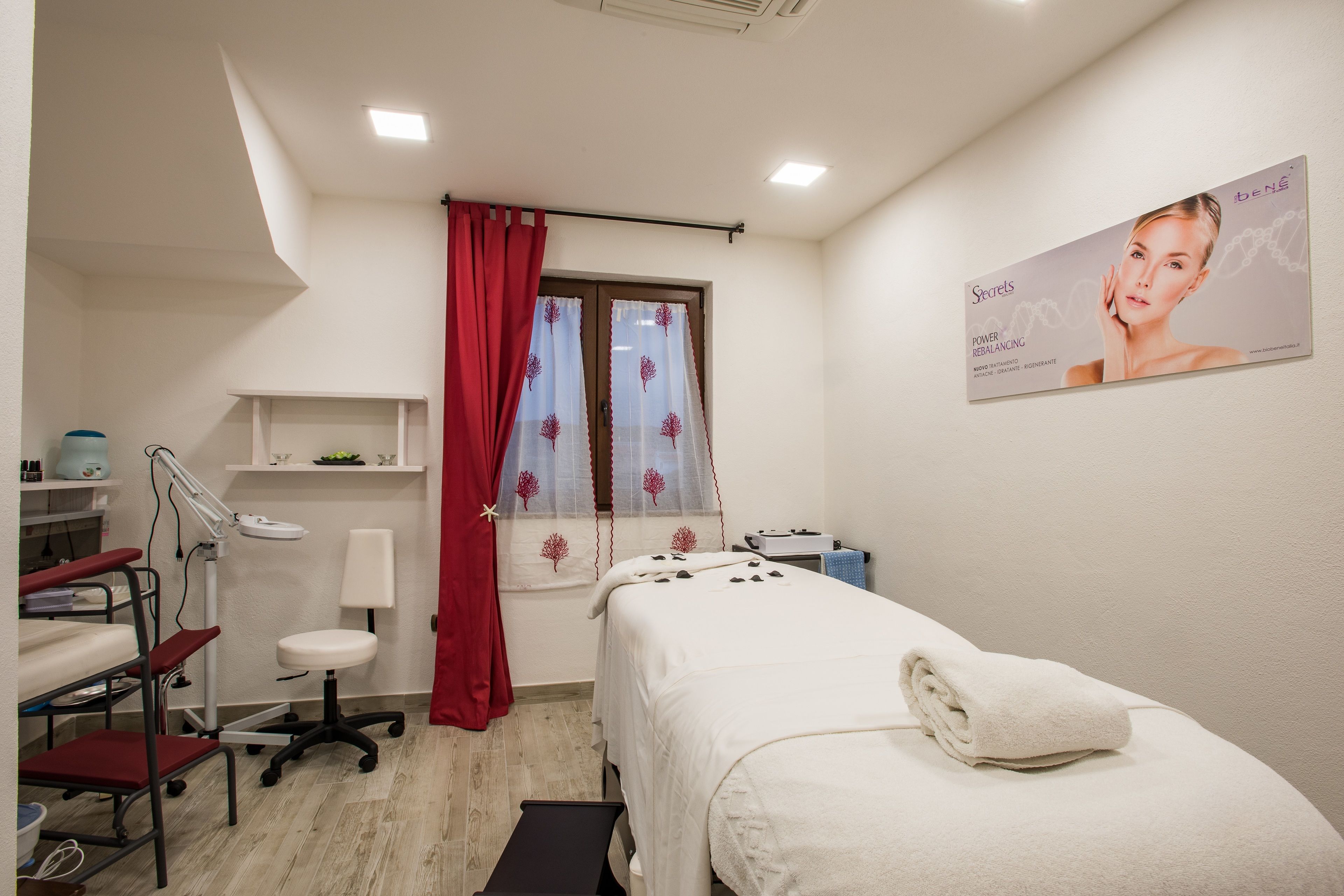 A clean, well-lit room with a bed, a desk, and a chair. There is a window with red curtains and a poster on the wall.