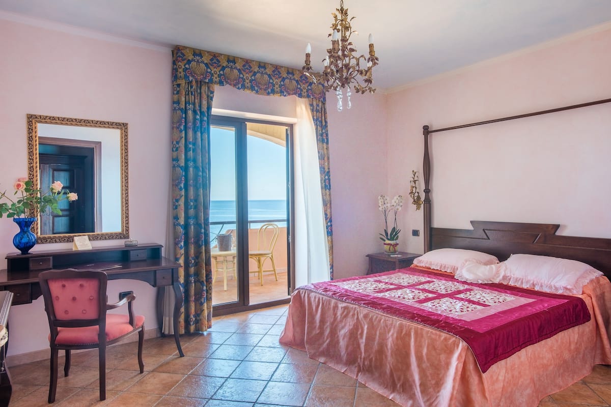 Ein Schlafzimmer mit Bett, Schreibtisch, Stuhl, Kommode, Spiegel, einer Kandelaber und einem Balkon mit Blick.