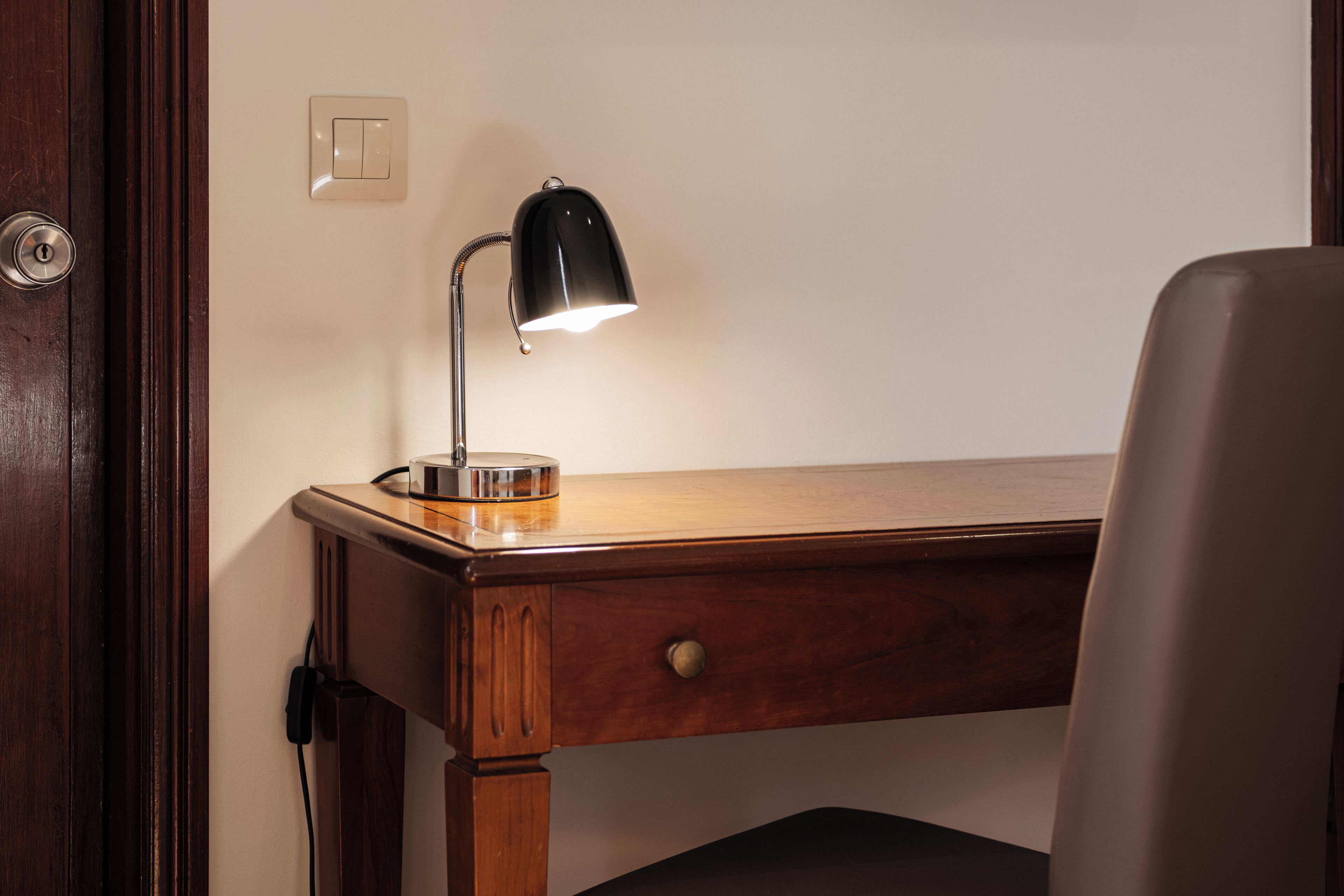 Una scrivania in legno con una lampada da scrivania nera, un cassetto e una presa di corrente.