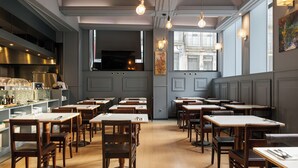 L’intérieur d’un restaurant, avec plusieurs tables et chaises, un espace cuisine et une grande fenêtre donnant sur la rue.