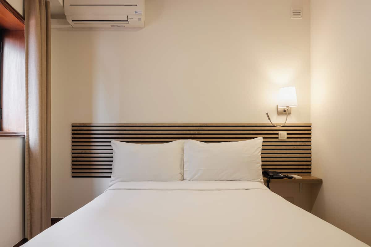 A hotel room with a bed, a nightstand, a wall-mounted lamp, and an air conditioning unit.