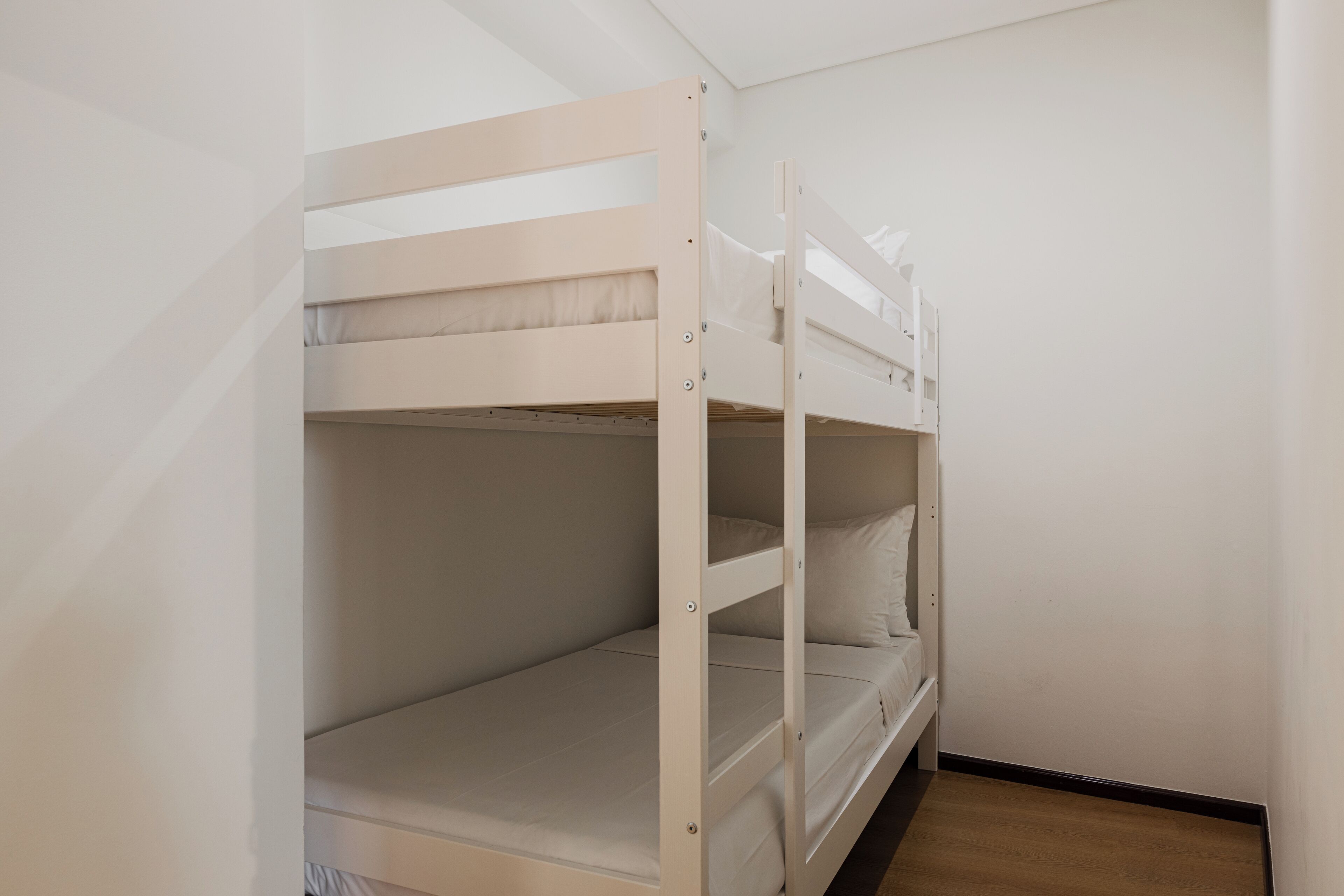 A bunk bed with a wooden frame and white bedding in a room with white walls and a wooden floor.