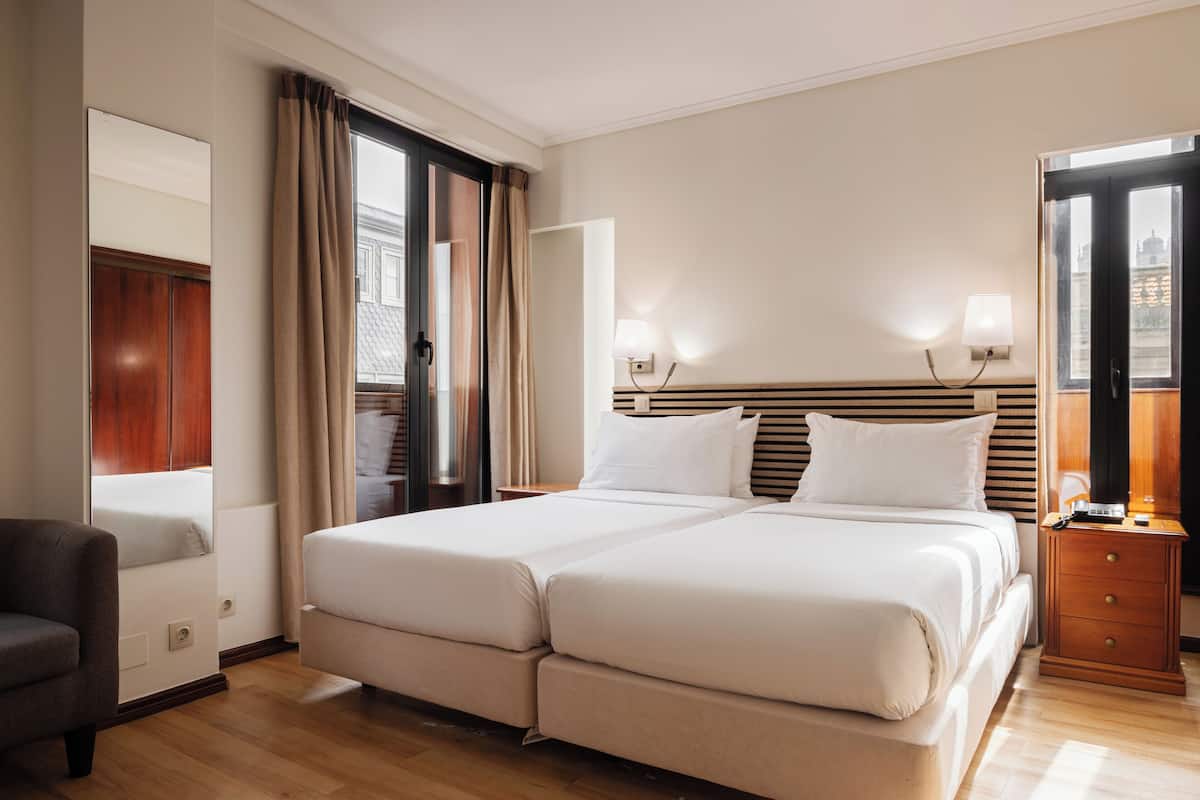 A hotel room with a double bed, a wooden nightstand, a lamp, a mirror, and a view of the city through the window.