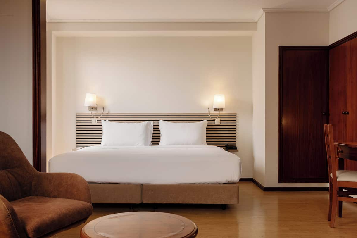 A hotel room with a large bed, two bedside lamps, a wooden chair, and a wooden table.