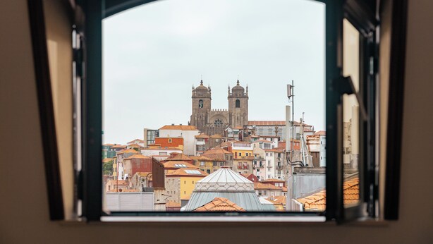 Une vue à travers la fenêtre d’un immeuble, sur une église imposante dotée de deux tours et d’un dôme, entourée de divers bâtiments aux couleurs de toits différentes.