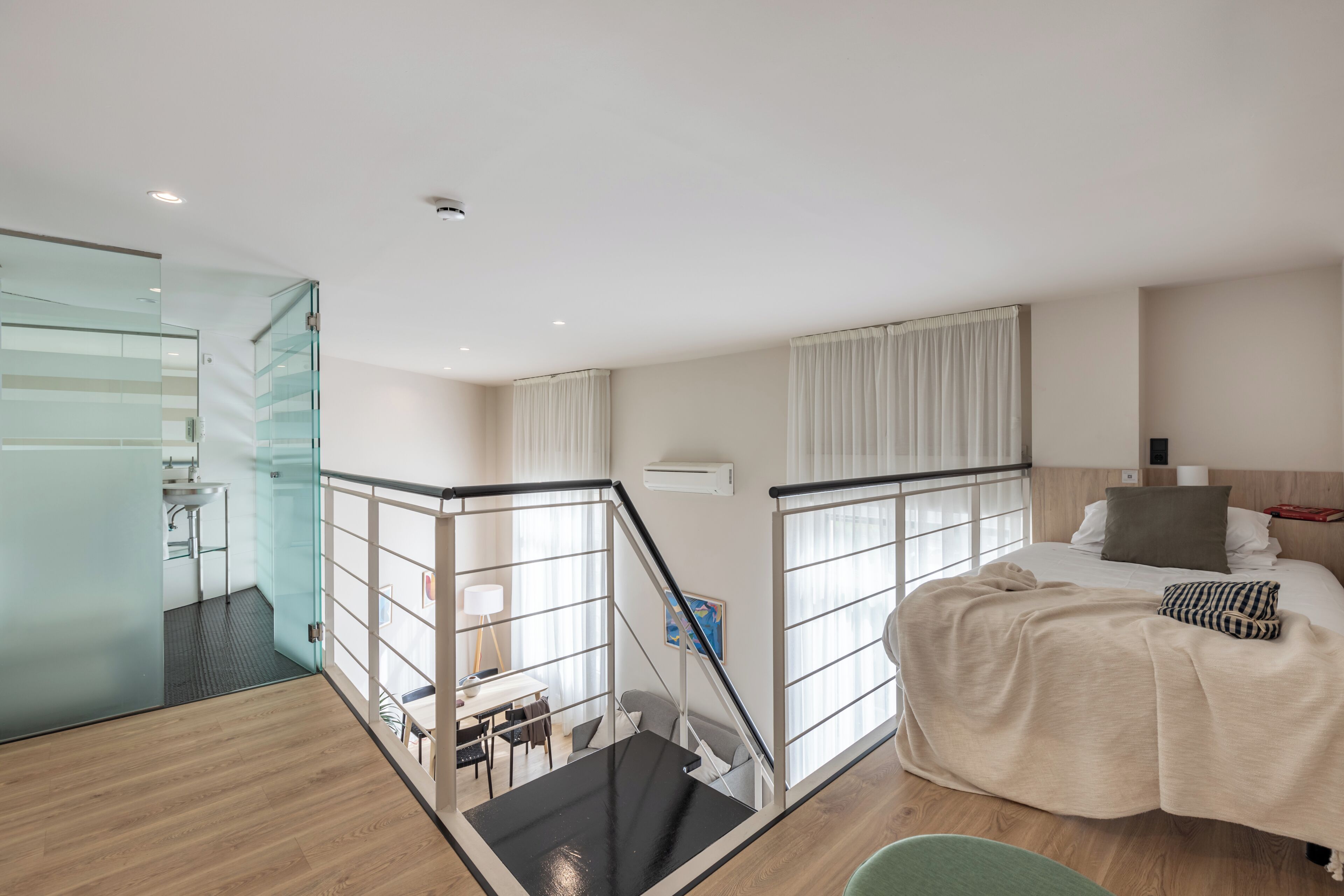 A modern hotel room with a glass partition, a staircase, a bed with a beige blanket, and a view of the city.