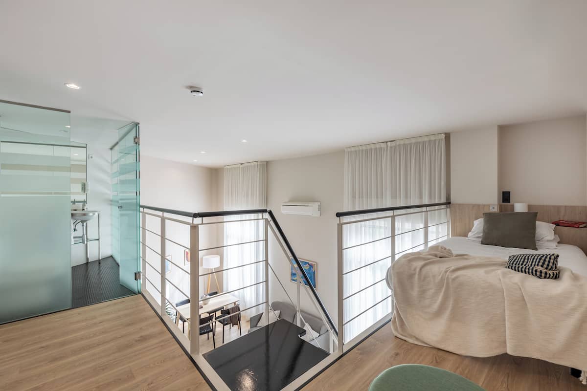 A modern hotel room with a glass partition, a staircase, a bed with a beige blanket, and a view of the city.