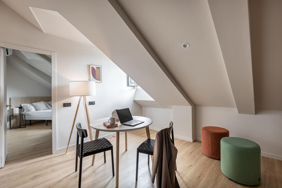 A modern loft bedroom with a sloped ceiling, a round dining table, two chairs, a laptop, and a green ottoman.