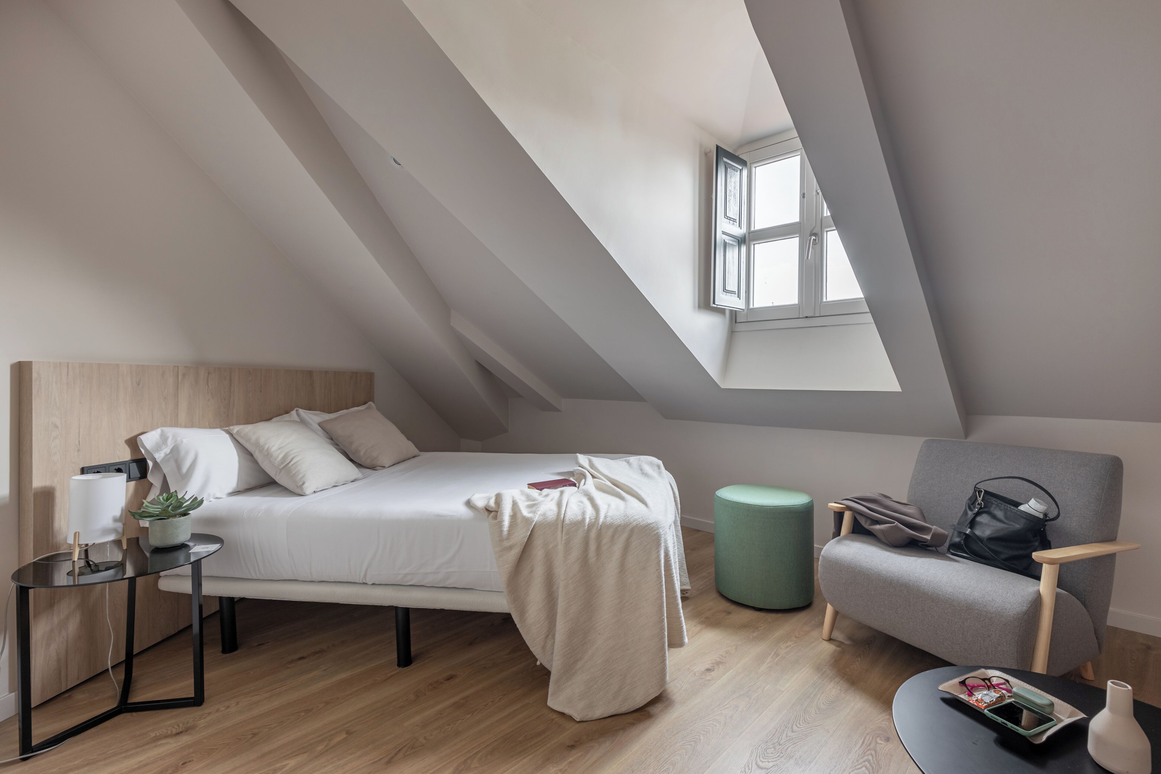 A modern loft bedroom with a sloped ceiling, a bed, a chair, a small table, and a window.