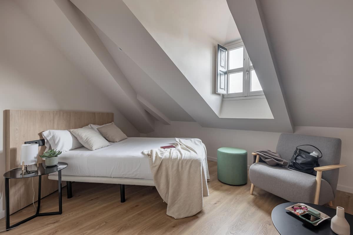 A modern loft bedroom with a sloped ceiling, a bed, a chair, a small table, and a window.