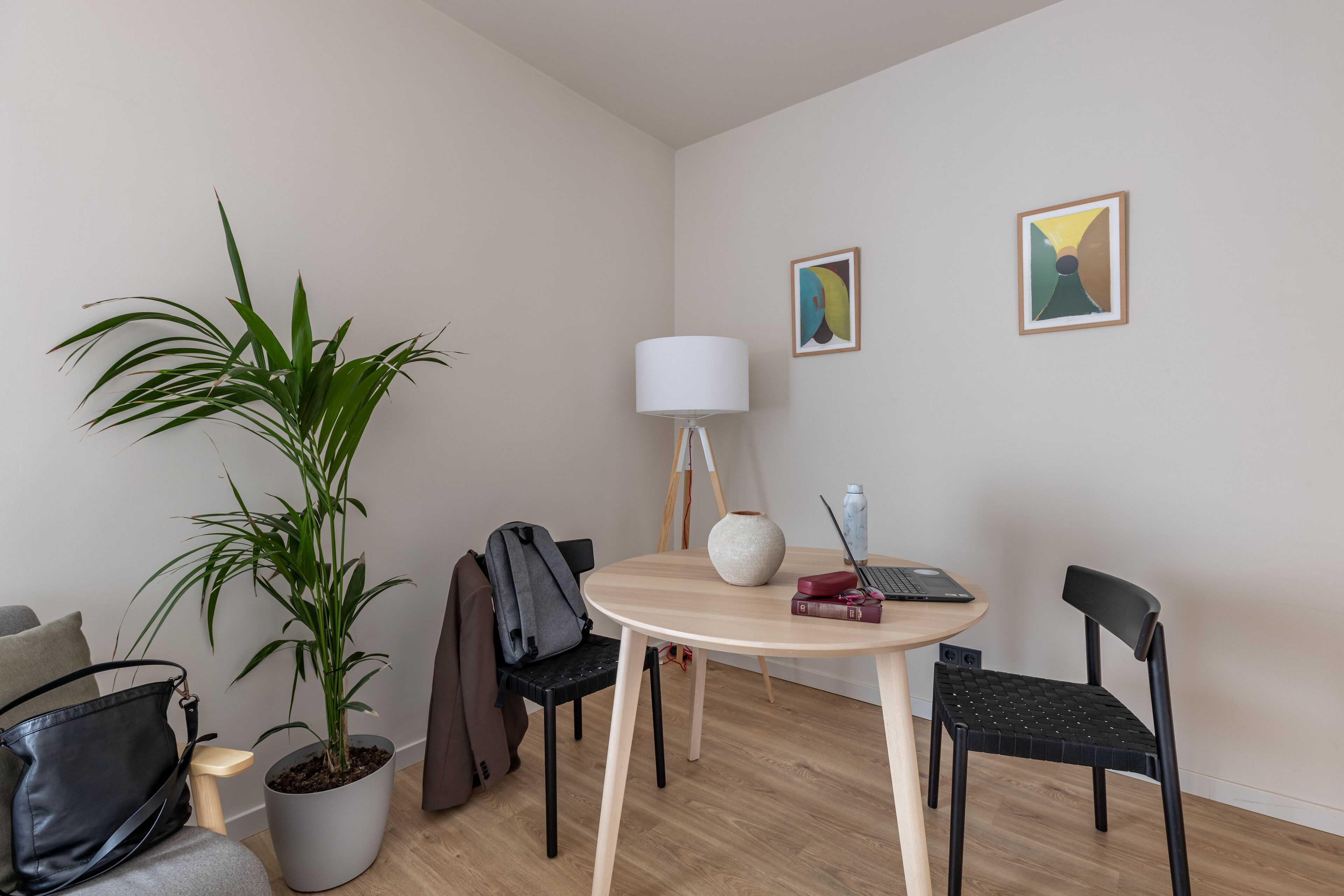 Une pièce moderne avec une table ronde en bois, deux chaises noires, une plante en pot, une lampe et deux œuvres d’art encadrées accrochées au mur.