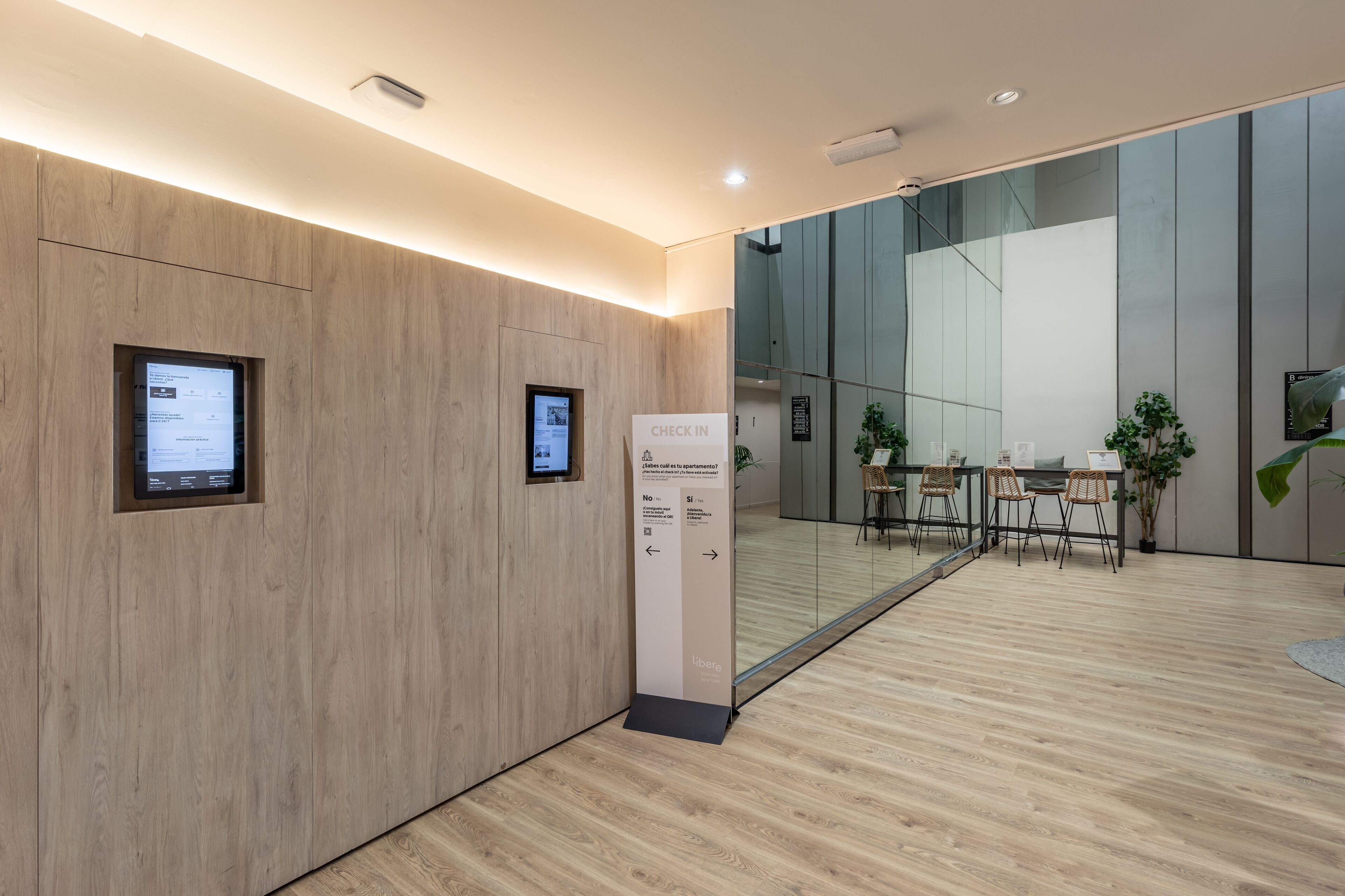 A modern interior with wooden panel walls, a check-in kiosk, and a glass partition leading to a seating area with wicker chairs and a table.