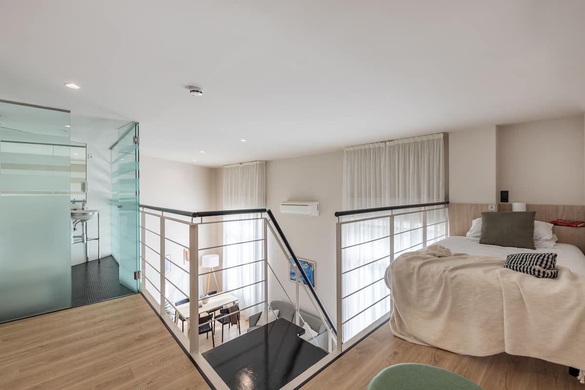 A modern hotel room with a glass partition, a staircase, a bed with a beige blanket, and a view of the city.