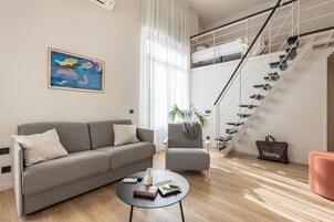 Una sala moderna con un sofá gris, una mesita redonda, una escalera con barandilla metálica y un ventanal con cortinas translúcidas.