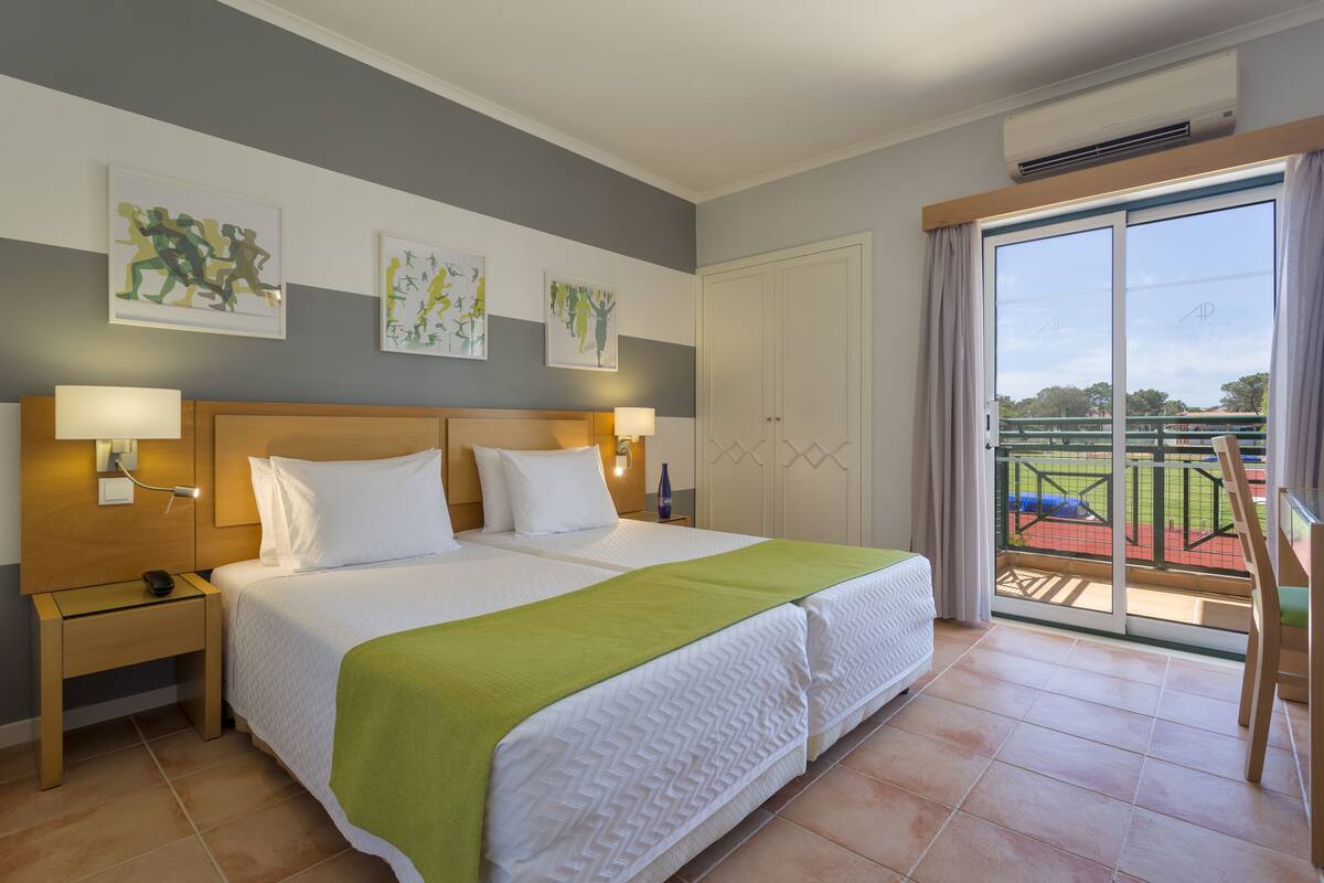 A hotel room with a large bed, two bedside lamps, a wooden nightstand, a green throw blanket, and a view of a tennis court through a sliding glass door.