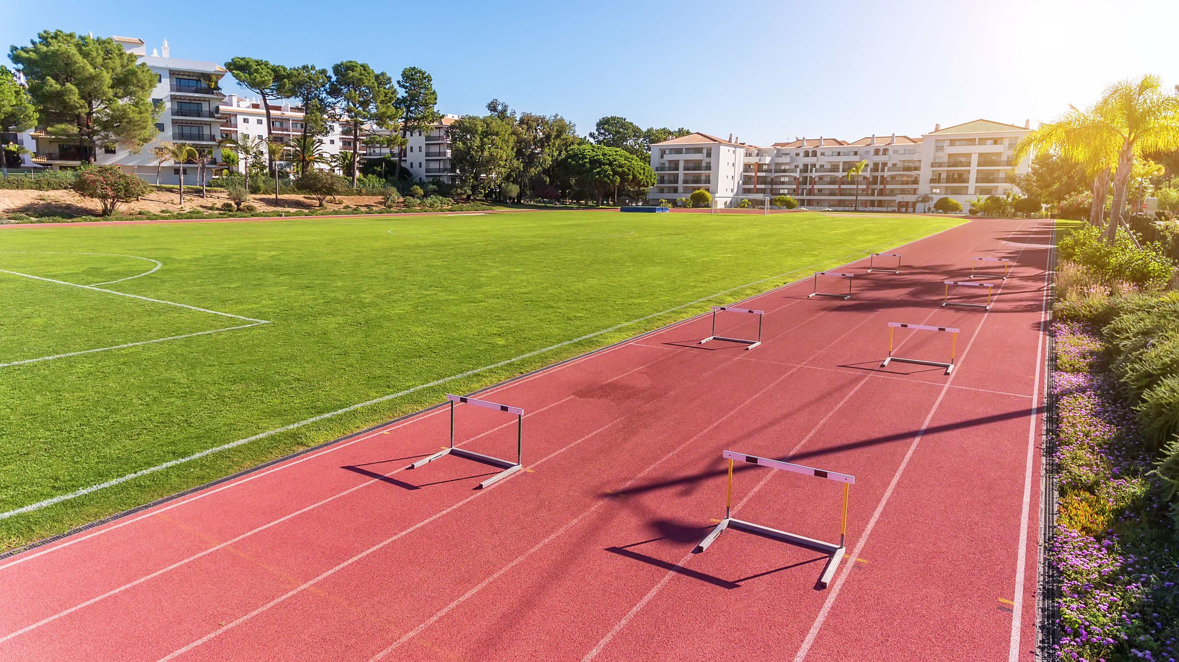 A red running track with hurdles, a green field, and buildings in the background.