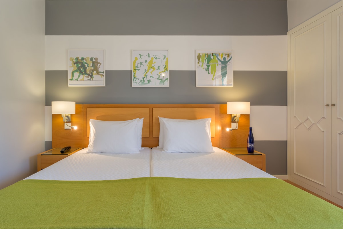 A double bed with white linens, two bedside tables with lamps, and three framed artworks on the wall.