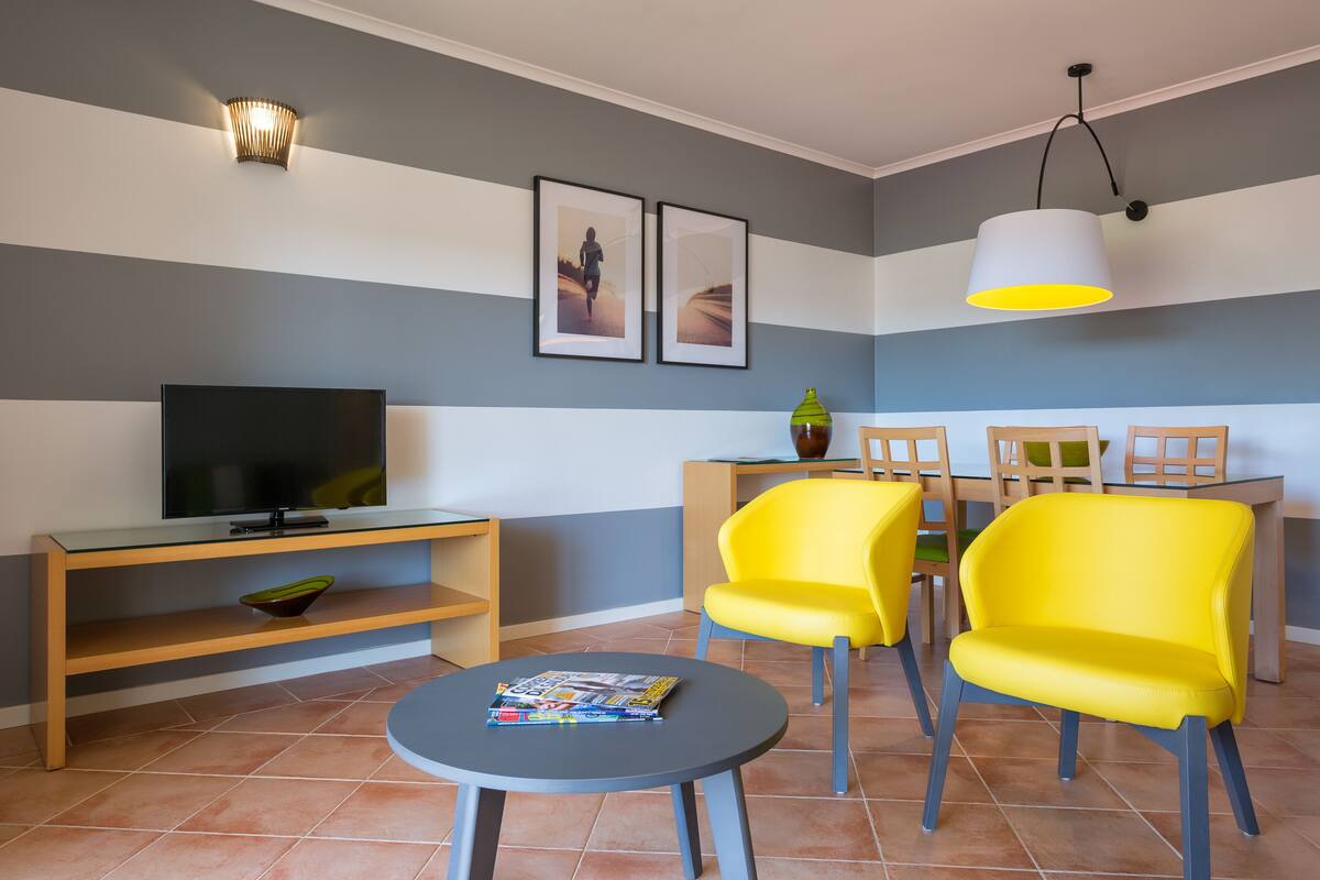 A modern living room with a flat-screen TV, a round coffee table, and two bright yellow chairs.