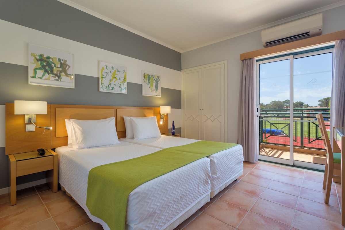 A hotel room with a large bed, two bedside lamps, a wooden nightstand, a green throw blanket, and a view of a tennis court through a sliding glass door.