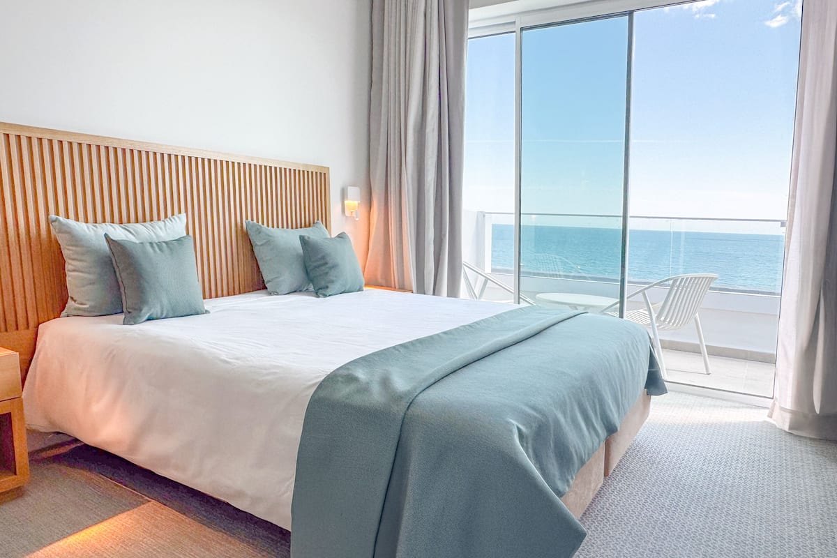A hotel room with a large bed, a wooden headboard, and a view of the sea through a balcony.