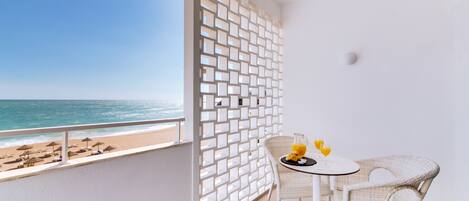 Un balcon avec vue sur la plage et l’océan, une table en osier blanc sur laquelle se trouve un verre de jus d’orange, et une chaise blanche.
