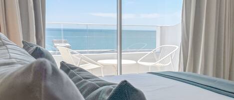 Quarto de hotel com varanda e vista para o mar, com cama, almofadas, uma pequena mesa e cadeiras brancas.