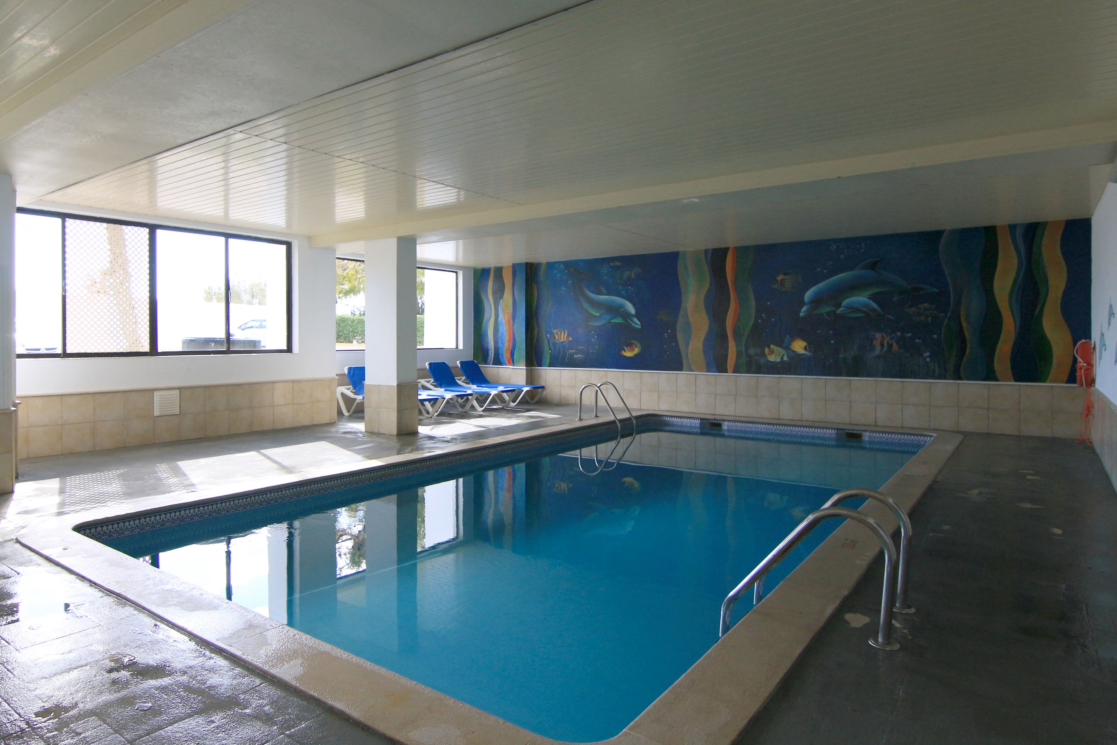 An indoor swimming pool with clear blue water, a tiled floor, and a mural of marine life on the wall.