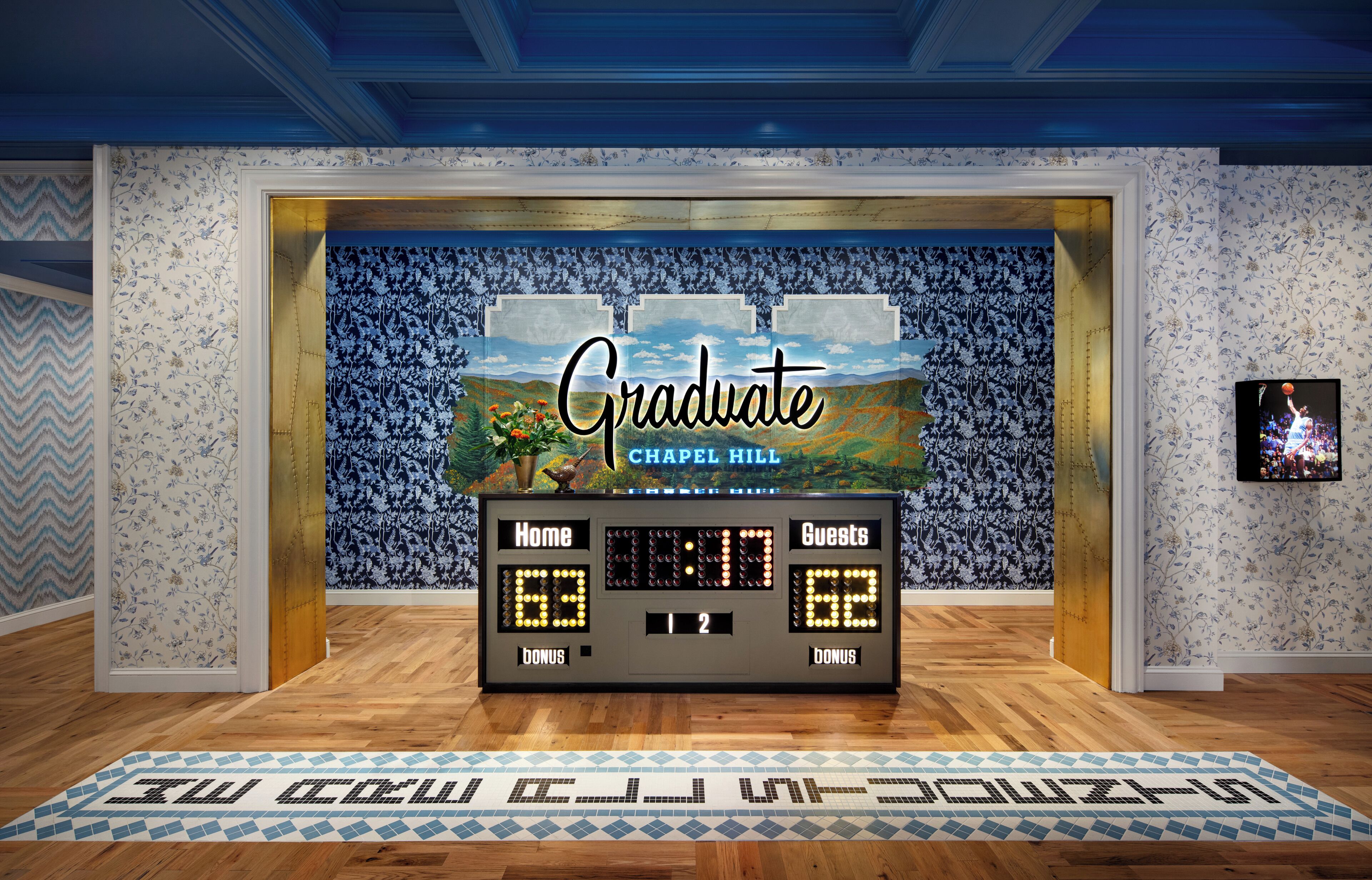 A scoreboard with 'Graduate Chapel Hill' and 'Home 63, Guests 62' displayed, surrounded by patterned wallpaper and a wooden floor.