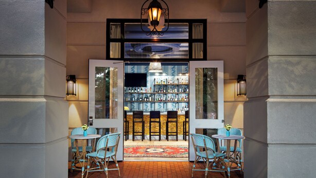 A hotel entrance with a blue awning, two chairs, a small table, and a bar area with bottles.