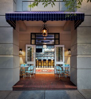 Entrada de un hotel con toldo azul, dos sillas, una mesa pequeña y una zona de bar con botellas.