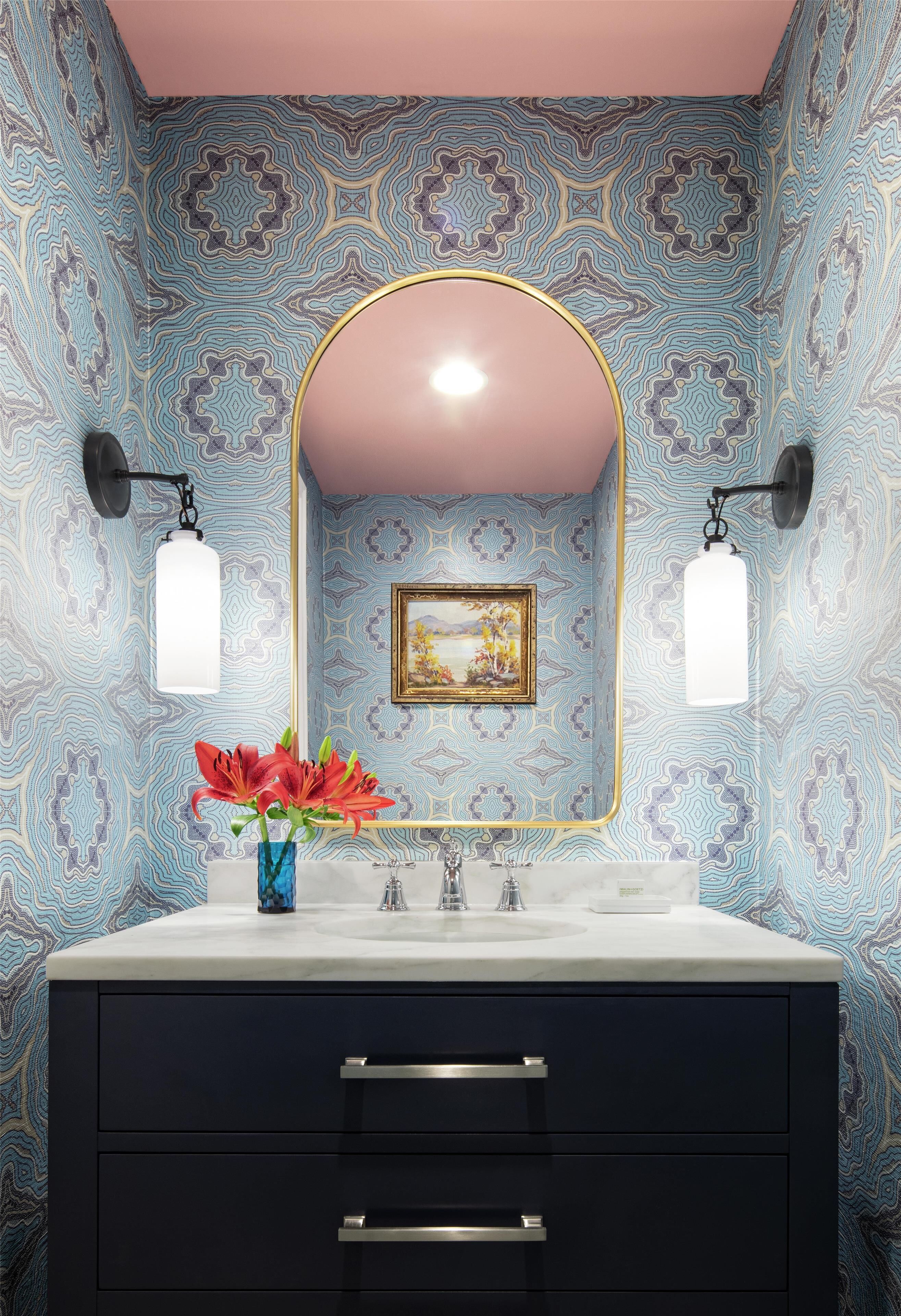 Une salle de bain avec un meuble-lavabo, deux appliques murales, un miroir avec un cadre doré et un vase rempli de fleurs rouges.