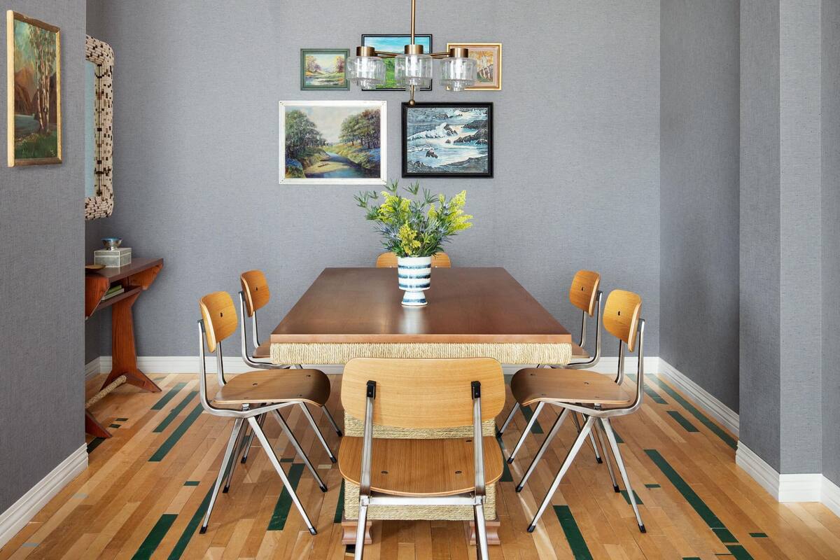 A dining area with a wooden table, four chairs, a vase with flowers, and framed artwork on the wall.
